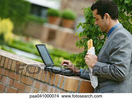 Picture - Businessman using laptop computer while eating sandwich outside. Fotosearch