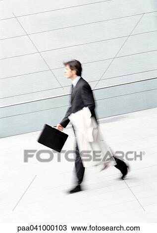 Stock Image - Businessman walking, holding overcoat and briefcase, blurred. Fotosearch