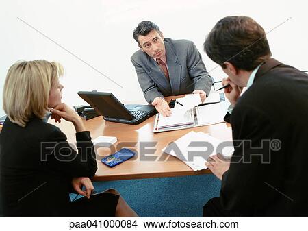 Picture - Two businessmen and a businesswoman sitting at desk, examining documents. Fotosearch