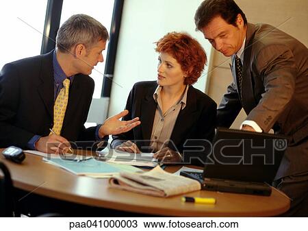 Stock Image - Two businessmen and a businesswoman around table, one using laptop computer. Fotosearch