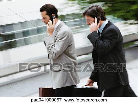 Two businessmen holding briefcase and cell phone, walking outside View Large Photo Image Stock Photo - Two businessmen holding briefcase and cell phone, walking outside. Fotosearch