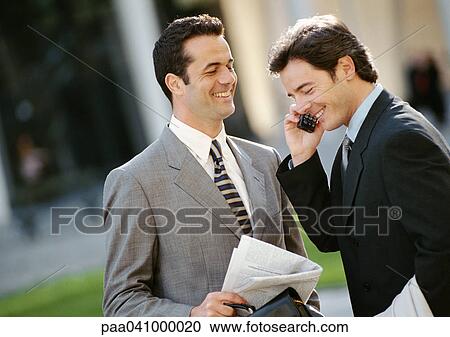 Stock Image - Two businessmen side by side outside, one using cell phone. Fotosearch
