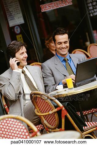 Stock Photo - Two businessmen sitting at cafe terrace, using cell phone and laptop computer. Fotosearch