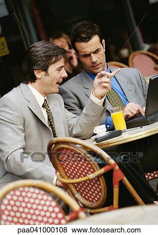 Stock Photo - Two businessmen sitting at cafe terrace, looking at laptop computer. Fotosearch