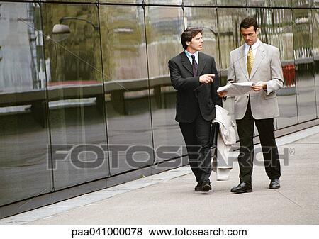 Two businessmen walking along sidewalk in front of building View Large Photo Image Stock Photo - Two businessmen walking along sidewalk in front of building. Fotosearch