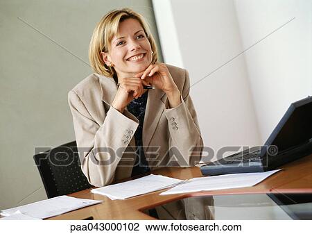 Stock Image - Businesswoman sitting at desk, hands under chin, smiling. Fotosearch
