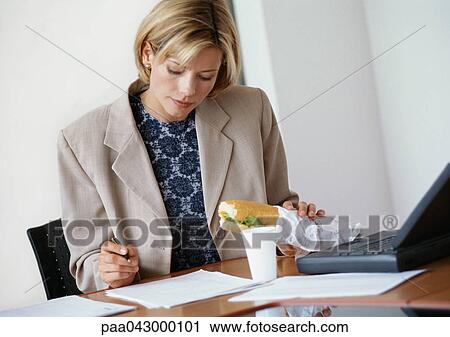 Stock Image - Businesswoman sitting at desk, holding sandwich and looking down at document. Fotosearch