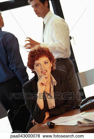Businesswoman sitting at desk, two businessmen talking in background View Large Photo Image Stock Photo - Businesswoman sitting at desk, two businessmen talking in background. Fotosearch