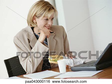 Stock Photograph - Businesswoman sitting at desk with laptop computer, holding sandwich. Fotosearch