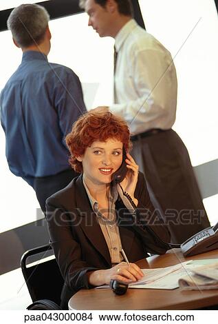 Businesswoman sitting, using phone, two businessmen in background View Large Photo Image Picture - Businesswoman sitting, using phone, two businessmen in background. Fotosearch