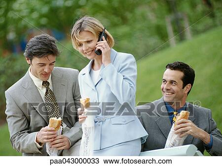 Three business people holding sandwiches outdoors, one using cell phone View Large Photo Image Stock Image - Three business people holding sandwiches outdoors, one using cell phone. Fotosearch
