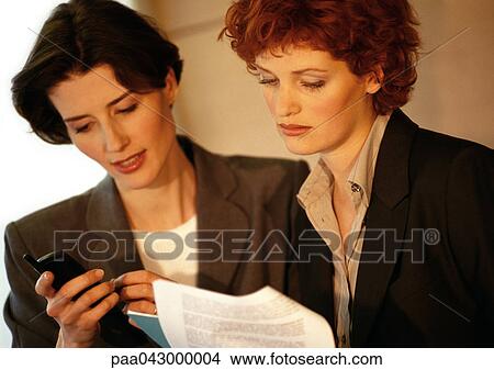 Two businesswomen side by side, one looking at cell phone View Large Photo Image Picture - Two businesswomen side by side, one looking at cell phone. Fotosearch