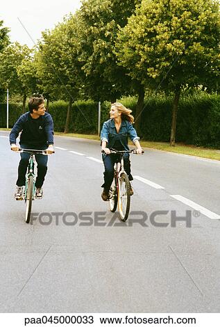Stock Image - Man and woman riding bikes side by side, looking at each other, full length, front view. Fotosearch