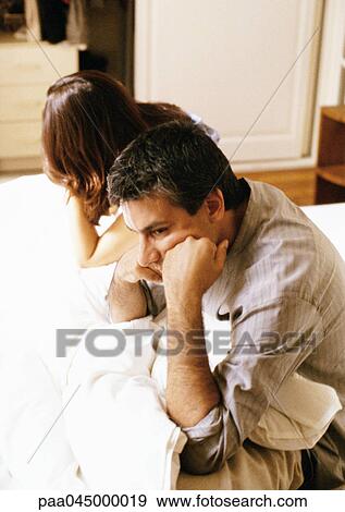 Stock Photo - Man and woman sitting up in bed, leaning heads on hands, waist up, side view. Fotosearch