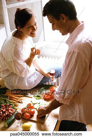 Stock Photography - Man chopping vegetables on table, woman sitting on table, holding glass. Fotosearch