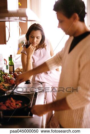Stock Image - Man cooking in blurred foreground, woman holding glass in background. Fotosearch
