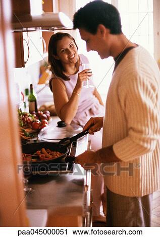 Stock Image - Man cooking, side view, woman holding glass, smiling. Fotosearch