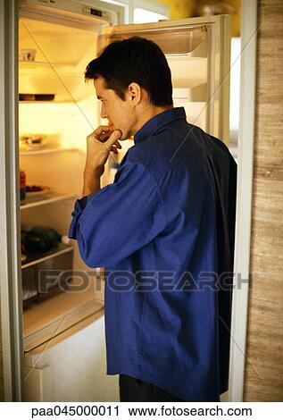 Stock Image - Man standing in front of open refrigerator, side view. Fotosearch