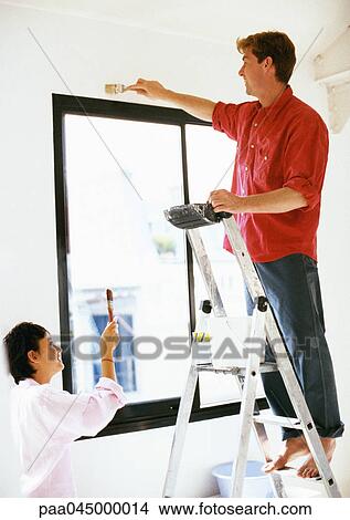 Picture - Man standing on ladder, painting wall, woman holding up brush. Fotosearch