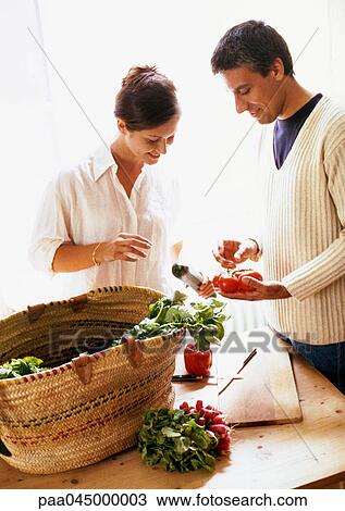 Stock Image - Woman and man standing behind market basket, looking at vegetables. Fotosearch