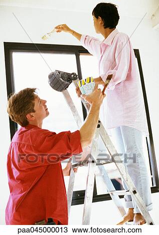 Stock Image - Woman painting wall, man holding ladder and paint can. Fotosearch
