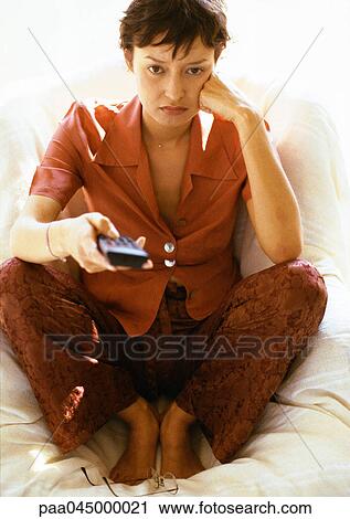 Stock Image - Woman sitting with knees up, leaning head on hand, holding remote control, full length, close-up. Fotosearch