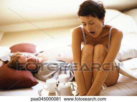 Stock Image - Woman sitting with knees up, yawning, beside man lying in bed. Fotosearch