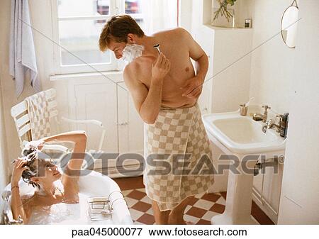 Stock Photo - Woman taking bath, man with towel wrapped around waist shaving, in bathroom. Fotosearch