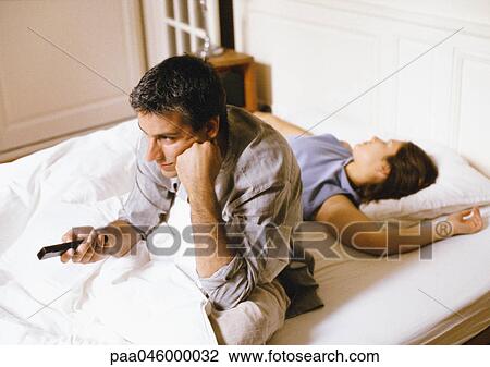 Man sitting up in bed, pointing remote control, woman lying behind him. View Large Photo Image Stock Image - Man sitting up in bed, pointing remote control, woman lying behind him.. Fotosearch
