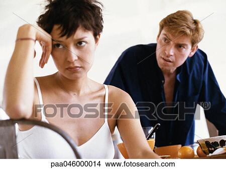 Woman sitting, elbow on back of chair, hand against head, man leaning toward her in background, front view View Large Photo Image Picture - Woman sitting, elbow on back of chair, hand against head, man leaning toward her in background, front view. Fotosearch