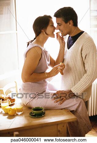 Stock Image - Woman sitting on table, feeding croissant to man. Fotosearch