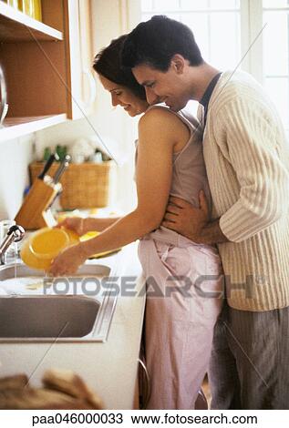 Stock Image - Woman washing dishes, man holding her waist and looking over her shoulder. Fotosearch