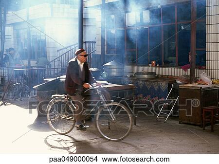 Picture - China, Xinjiang Province, Turpan, man riding bicycle near shop front. Fotosearch