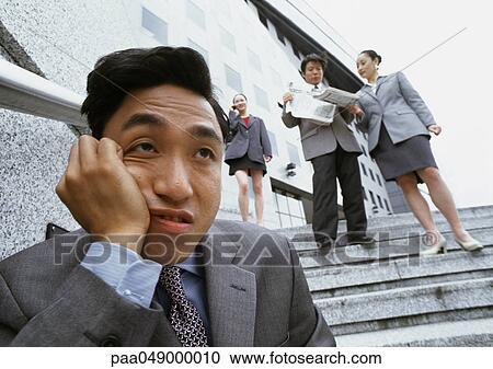 Stock Image - Man sitting on stairs, hand on cheek, people in background. Fotosearch