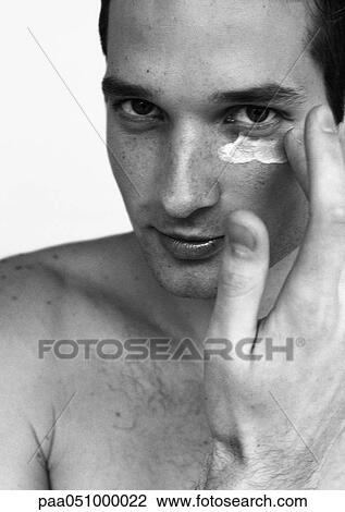 Stock Image - Man putting cream under eye, looking at camera, portrait, b&w. Fotosearch
