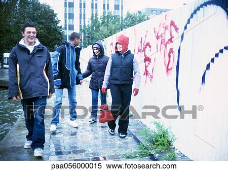 Stock Photography - Four young people walking on sidewalk next to graffiti-covered wall. Fotosearch