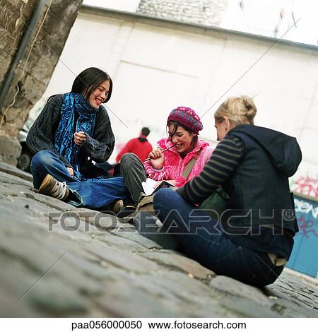 Stock Image - Three teenage girls sitting on cobblestones, smiling. Fotosearch