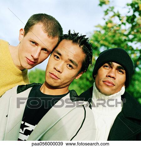 Stock Photo - Three young men standing together, head and shoulders, tilt, trees and sky in background. Fotosearch