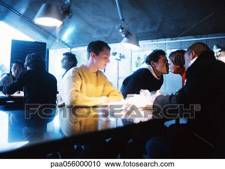 Stock Image - Young people sitting at table, couple kissing. Fotosearch