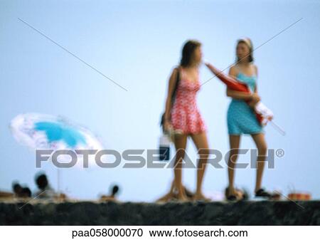 Stock Image - Two women walking on beach, one holding parasol. Fotosearch