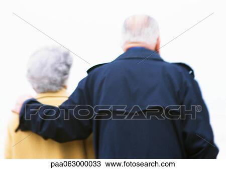 Stock Image - Elderly man and woman, man's arm around woman's shoulder, rear view, blurred and backlit. Fotosearch