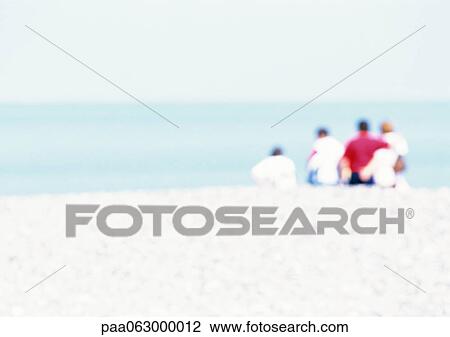 Stock Image - Group of people sitting on sand at the beach, rear view, blurred. Fotosearch