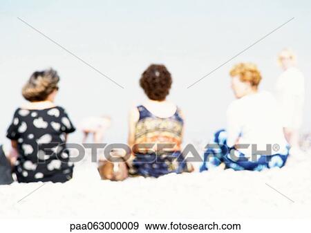 Stock Photo - People sitting on sand at the beach, rear view, blurred. Fotosearch