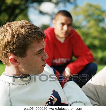 Stock Image - Close up of young man sitting outside, second young man crouching in background, blurred.. Fotosearch