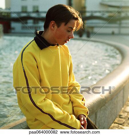 Stock Image - Teenage boy sitting on edge of fountain, looking down.. Fotosearch