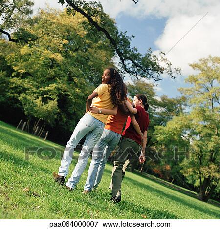 Stock Photo - Three young women walking on grass with arms around each other, rear view, one looking over shoulder. Fotosearch