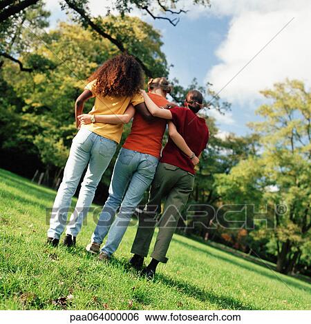 Stock Photograph - Three young women walking on grass with arms around each other, rear view, full length. Fotosearch