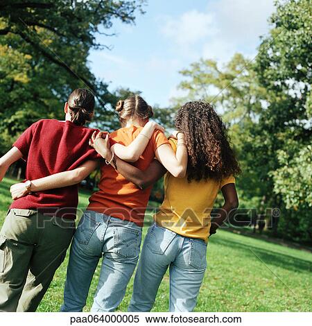 Stock Photography - Three young women walking on grass with arms around each other, rear view. Fotosearch