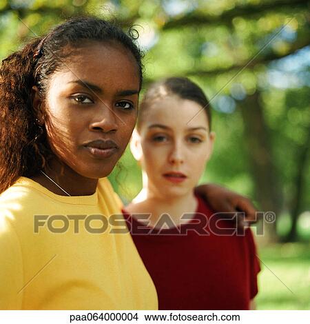 Two young women standing together outside, one with arm around other's shoulder, looking at camera View Large Photo Image Picture - Two young women standing together outside, one with arm around other's shoulder, looking at camera. Fotosearch