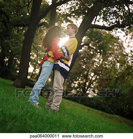 Stock Image - Young man and woman standing on grass, embracing, side view, full length. Fotosearch
